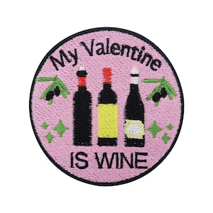 Pode incluir: Etiqueta redonda bordada rosa com o texto "My Valentine is Wine" e três garrafas de vinho, duas vermelhas e uma branca, com ramos de oliveira verdes de cada lado.