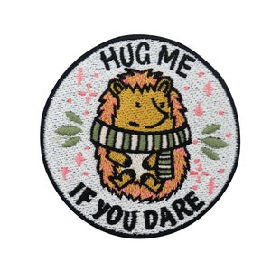 May include: Embroidered patch with a white background and black outline. The patch features a cartoon hedgehog wearing a green and white striped scarf. The text "Hug Me If You Dare" is written in a black cursive font around the hedgehog.