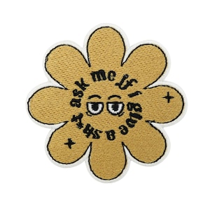 Puede incluir: Parche bordado con forma de flor, con centro dorado y contorno blanco. El parche presenta el texto "ask me if I give a sh*t" en un círculo alrededor de unos ojos de dibujos animados.