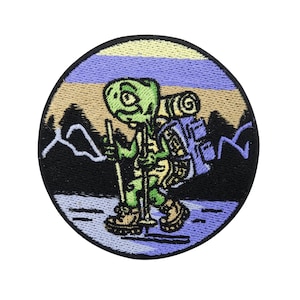 Può includere: Una toppa ricamata con una tartaruga dei cartoni animati che indossa uno zaino e fa escursionismo con un bastone da trekking. La tartaruga è verde, gialla e marrone. Lo sfondo è un tramonto blu, viola e marrone con montagne.