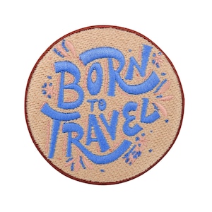 Peut inclure: Un patch rond brodé beige avec une bordure marron. Le patch présente le texte "Born to Travel" en broderie bleue avec des accents floraux roses.