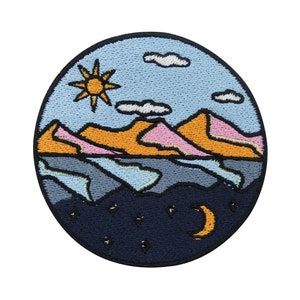 Könnte beinhalten: Gestickter Aufnäher mit einem runden Design, das einen blauen Himmel mit Wolken, orangefarbene und rosafarbene Berge, eine blaue Wasserfläche und einen dunkelblauen Nachthimmel mit Sternen und einer Mondsichel zeigt.