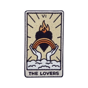Op de afbeelding: Een beige geborduurde patch met een zwart hart, een regenboog en twee handen die de regenboog vasthouden. De tekst "VI THE LOVERS" staat onderaan de patch.