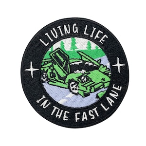 May include: A black and white embroidered patch featuring a green car and the text "Living Life In The Fast Lane".
