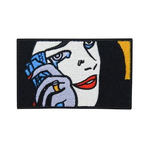 Op de afbeelding: Geborduurde patch met een vrouw met een traan die over haar wang rolt, een stapel geld in de ene hand en een ring in de andere. De achtergrond is zwart met een gele rand.
