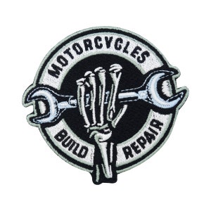 Puede incluir: Un parche bordado en blanco y negro con una mano de esqueleto sosteniendo una llave inglesa. El parche está rodeado por un círculo blanco con el texto "BUILD MOTORCYCLES REPAIR" en negro.