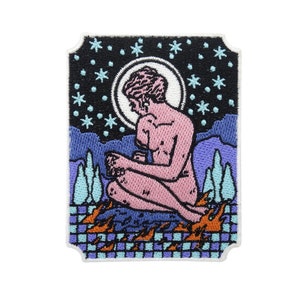 Puede incluir: Parche bordado con una figura rosa sentada con una luna y estrellas en el fondo. La figura está rodeada de llamas y un patrón de cuadros.