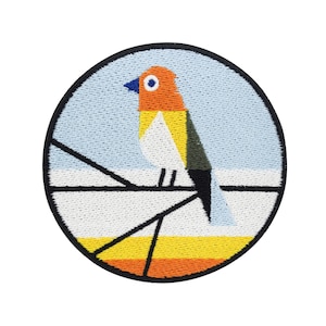 May include: A round embroidered patch featuring a stylised bird in orange, yellow, white and green perched on a branch. The background is blue.