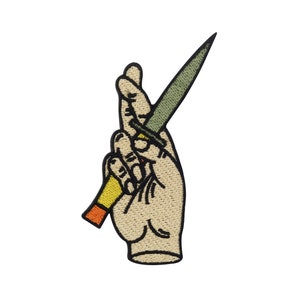 Op de afbeelding: Een geborduurde patch van een hand die een mes vasthoudt met een geel en oranje handvat. Het mes is groen en de hand is beige.