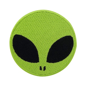 May include: Green alien face embroidered patch with black eyes. The patch is circular and has a stitched border.