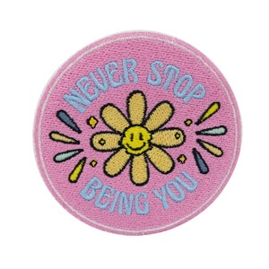 May include: Pink round embroidered patch with a yellow flower with a smiley face in the center. The text "NEVER STOP BEING YOU" is embroidered around the flower in blue.