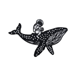 Peut inclure: Patch brodé noir et blanc d'un astronaute chevauchant une baleine avec un motif étoilé.