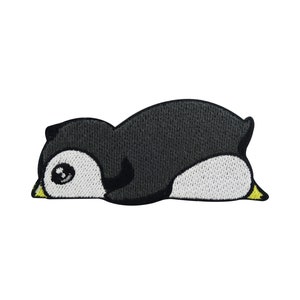 May include: Embroidered iron-on patch of a black and white penguin lying on its belly with its head turned to the side. The penguin has a yellow beak and feet.