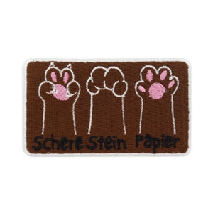May include: Embroidered patch with three cat paw designs in brown, white, and pink. The patch has a white border and the German text "Schere Stein Papier" is printed at the bottom. Ideal for customising clothing or accessories.