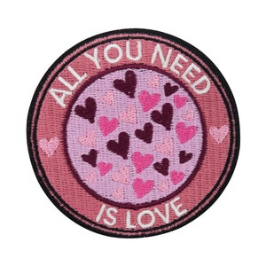 Puede incluir: Un parche bordado rosa y negro con el texto "All You Need Is Love" rodeado de un círculo de corazones rosas.