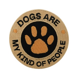 Może przedstawiać: Okrągła, beżowa łatka z czarnymi i pomarańczowymi haftami. Łatka zawiera napis "Dogs are my kind of people" z odciskiem łapy w środku.