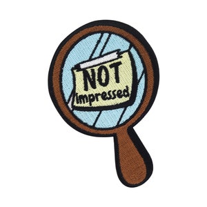 Può includere: Toppa ricamata con un design a forma di lente d'ingrandimento. La lente è azzurra e il manico marrone. All'interno della lente c'è un foglio giallo con le parole "NOT impressed" in nero.