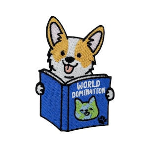 Könnte beinhalten: Gestickter Aufnäher mit einem lächelnden Corgi-Hund, der ein blaues Buch mit der Aufschrift "WORLD DOMINATION" liest. Das Buch hat eine grün-blaue Grafik einer Katze mit einem Pfotenabdruck. Der Corgi hat weißes, orangefarbenes und schwarzes Fell.