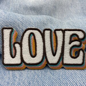 Puede incluir: Un parche bordado blanco con la palabra "LOVE" en estilo retro. Las letras est&aacute;n delineadas en marr&oacute;n, azul verdoso, rosa y naranja.