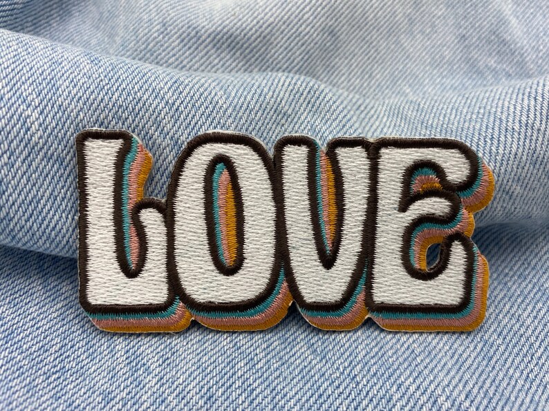 Puede incluir: Un parche bordado blanco con la palabra "LOVE" en estilo retro. Las letras est&aacute;n delineadas en marr&oacute;n, azul verdoso, rosa y naranja.