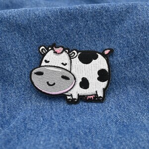 May include: Embroidered patch of a cartoon cow with a white body, black spots, and a pink inner ear. The cow has a grey face and a smiling expression. The patch is on a blue denim background.