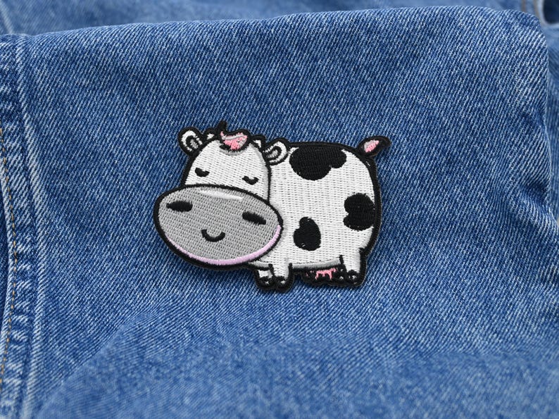 May include: Embroidered patch of a cartoon cow with a white body, black spots, and a pink inner ear. The cow has a grey face and a smiling expression. The patch is on a blue denim background.