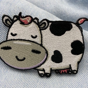 May include: Embroidered patch depicting a cartoon cow with a smiling face and closed eyes. The cow has white fur with black spots, a grey muzzle, and a pink flower. The patch is set against a denim background.