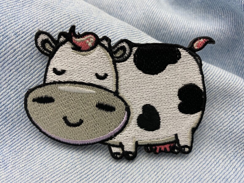 May include: Embroidered patch depicting a cartoon cow with a smiling face and closed eyes. The cow has white fur with black spots, a grey muzzle, and a pink flower. The patch is set against a denim background.