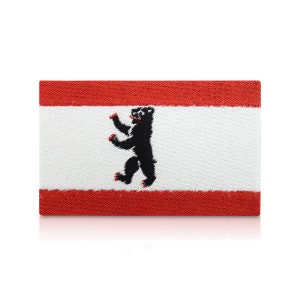 May include: A red and white embroidered patch with a black bear standing on its hind legs. The bear is the symbol of Berlin, Germany.
