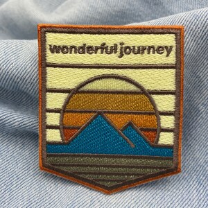 May include: A brown and orange embroidered patch with a sunset and mountains design. The text "wonderful journey" is embroidered in brown on the top of the patch.
