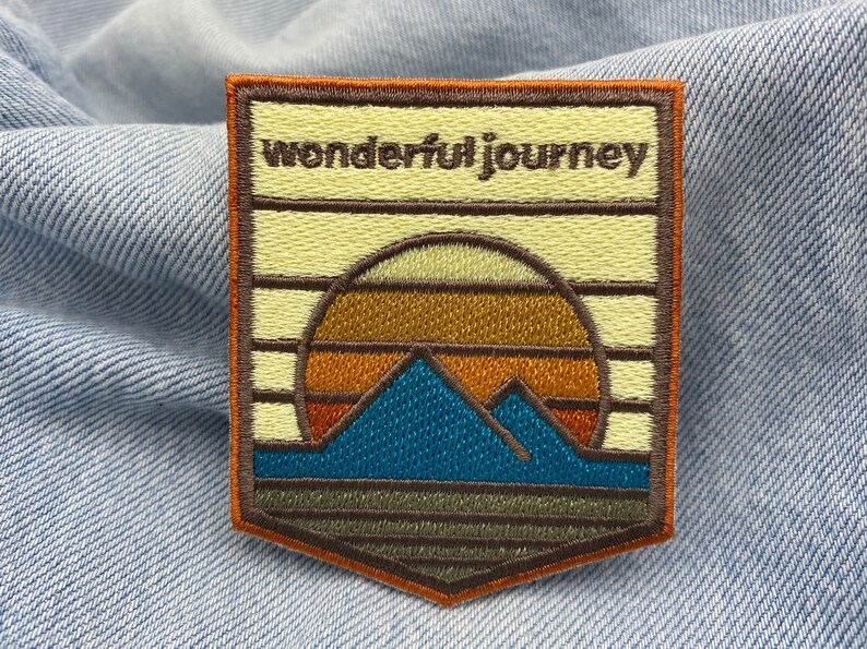 May include: A brown and orange embroidered patch with a sunset and mountains design. The text "wonderful journey" is embroidered in brown on the top of the patch.