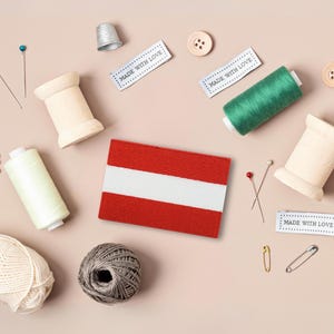 May include: A flat lay of sewing supplies, including spools of thread, buttons, pins, and a thimble. There is also a small fabric flag of Austria with the text "Made with Love" on three labels.