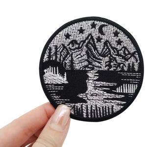 Puede incluir: Parche bordado en blanco y negro que representa una cordillera con una luna creciente y estrellas sobre un lago. El parche es circular y tiene un contorno blanco.