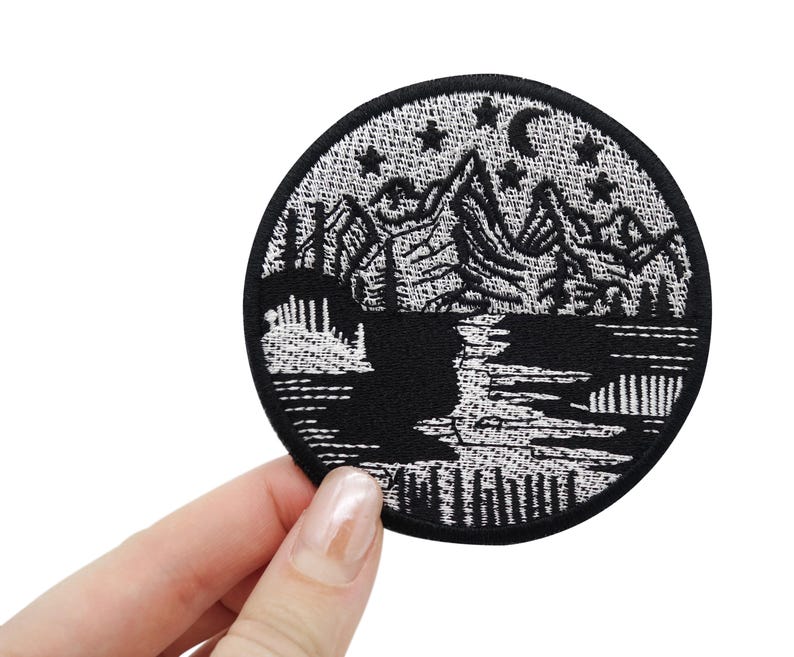 Puede incluir: Parche bordado en blanco y negro que representa una cordillera con una luna creciente y estrellas sobre un lago. El parche es circular y tiene un contorno blanco.