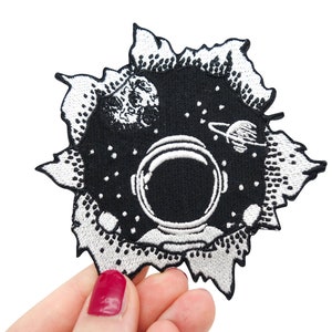 May include: Black and white embroidered patch of an astronaut's helmet in space with a planet and stars. The patch is designed to look like a hole in a fabric surface.