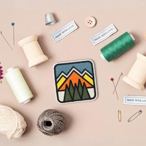 May include: A colorful embroidered patch with a mountain scene, surrounded by spools of thread, buttons, pins, and safety pins. The patch has the text "Made with Love" on three labels.