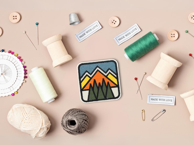 May include: A colorful embroidered patch with a mountain scene, surrounded by spools of thread, buttons, pins, and safety pins. The patch has the text "Made with Love" on three labels.