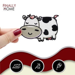 May include: Embroidered cow patch with a smiling face, black spots, and a pink tuft of hair. The patch is white with black spots and a light grey face. The text "FINALLY HOME" is in the upper left corner.