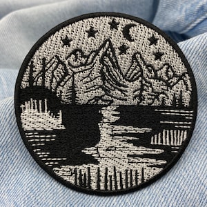 Puede incluir: Parche bordado en blanco y negro que representa una cordillera con un lago en primer plano. Las monta&ntilde;as est&aacute;n delineadas en blanco con detalles en negro. El lago es negro con olas blancas. Hay estrellas y una luna creciente en el cielo sobre las monta&ntilde;as.