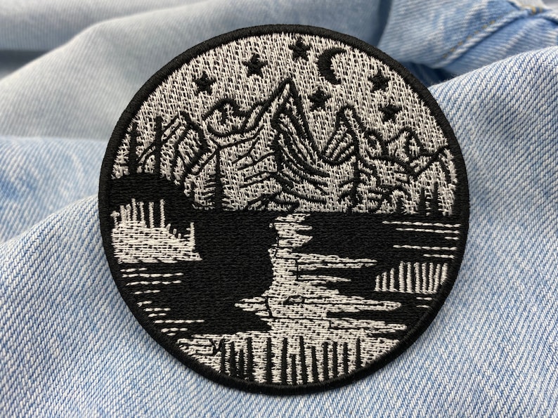 Puede incluir: Parche bordado en blanco y negro que representa una cordillera con un lago en primer plano. Las monta&ntilde;as est&aacute;n delineadas en blanco con detalles en negro. El lago es negro con olas blancas. Hay estrellas y una luna creciente en el cielo sobre las monta&ntilde;as.