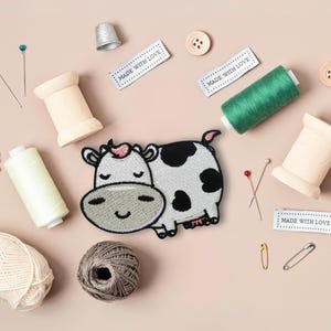 May include: A collection of sewing items on a pale pink background. Features a cow patch, thread spools, buttons, pins, yarn, a thimble, and labels with the text "MADE WITH LOVE."
