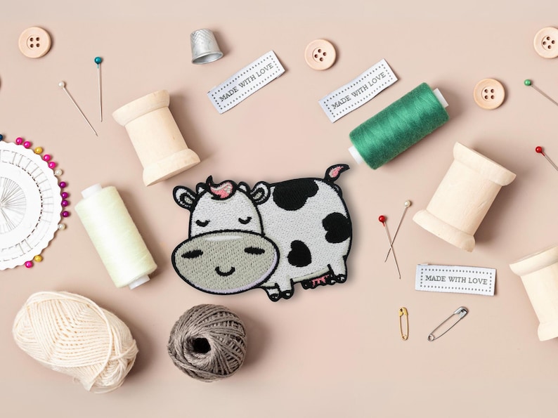 May include: A collection of sewing items on a pale pink background. Features a cow patch, thread spools, buttons, pins, yarn, a thimble, and labels with the text "MADE WITH LOVE."