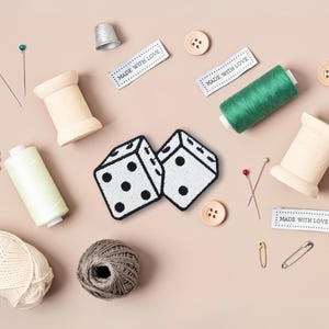 Pu&ograve; includere: Due dadi bianchi con punti neri, circondati da materiali per cucire, tra cui bobine di filo, spilli e aghi. L'immagine include anche etichette che recitano "Made with Love".