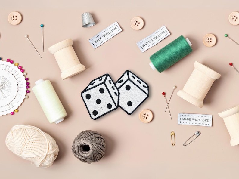 Pu&ograve; includere: Due dadi bianchi con punti neri, circondati da materiali per cucire, tra cui bobine di filo, spilli e aghi. L'immagine include anche etichette che recitano "Made with Love".