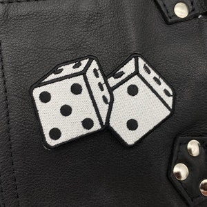 Pu&ograve; includere: Due dadi bianchi con punti neri cuciti su una superficie in pelle nera. I dadi si sovrappongono leggermente. La superficie presenta una cucitura e una fibbia in metallo.