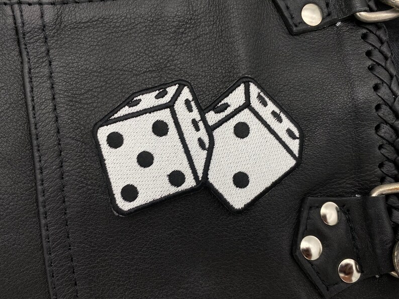 Pu&ograve; includere: Due dadi bianchi con punti neri cuciti su una superficie in pelle nera. I dadi si sovrappongono leggermente. La superficie presenta una cucitura e una fibbia in metallo.