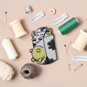 May include: An embroidered patch featuring a cartoon cow in a milk carton, with white and black stitching. The patch is surrounded by sewing supplies, including spools of thread, pins, and needles. The text "Made with Love" appears on three white labels.