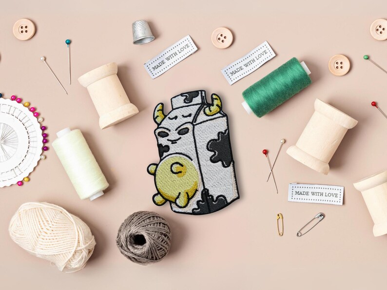 May include: An embroidered patch featuring a cartoon cow in a milk carton, with white and black stitching. The patch is surrounded by sewing supplies, including spools of thread, pins, and needles. The text "Made with Love" appears on three white labels.