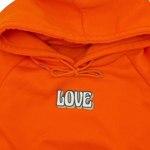 Puede incluir: Sudadera naranja con capucha con un gr&aacute;fico blanco de "LOVE" en la parte delantera. El gr&aacute;fico tiene un estilo retro, 3D con contornos azules y amarillos.