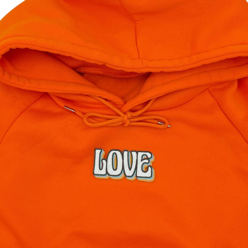 Puede incluir: Sudadera naranja con capucha con un gr&aacute;fico blanco de "LOVE" en la parte delantera. El gr&aacute;fico tiene un estilo retro, 3D con contornos azules y amarillos.